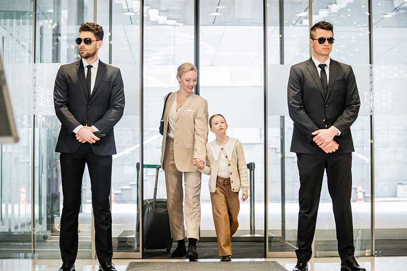 Private security concept, two bodyguards in formal wear and sunglasses standing near hotel entrance, cheerful woman and child holding hands and walking with luggage, entering lobby, luxury lifestyle