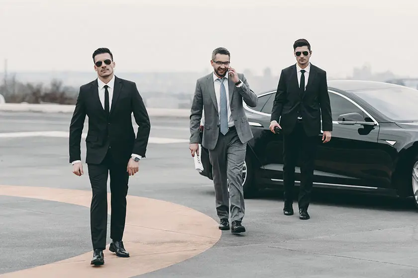 smiling-businessman-walking-bodyguards-car-talking-smartphone